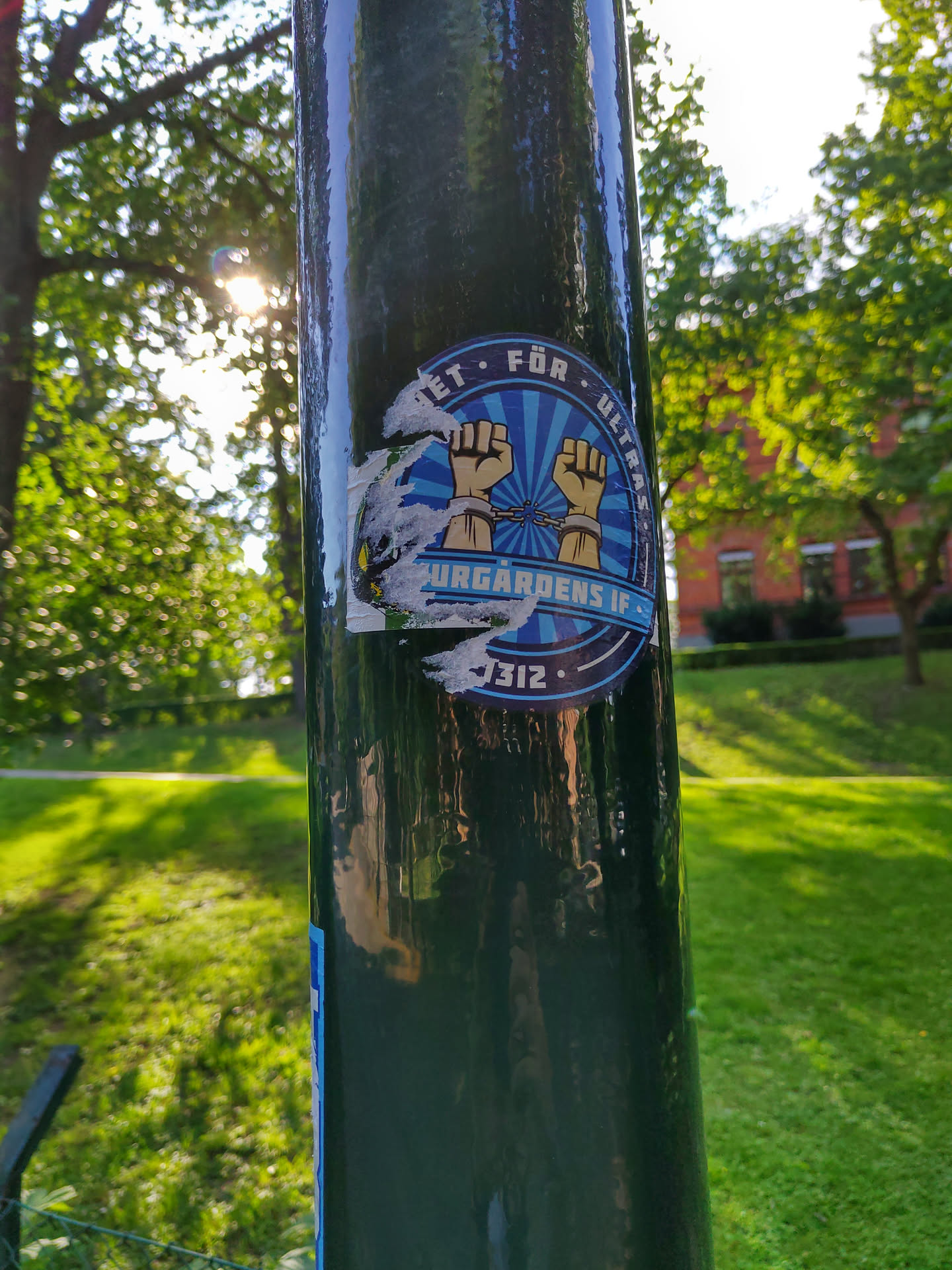 A round sticker affixed to a dark-green pole. The sticker features a blue circular design with two light-brown fists in chains, and text in a circular shape surrounding the fists.  Part of the sticker is damaged. The text partially visible is 'ET FÖR ULTRA' and 'Djurgårdens IF 1912'. The background is a green lawn and trees.