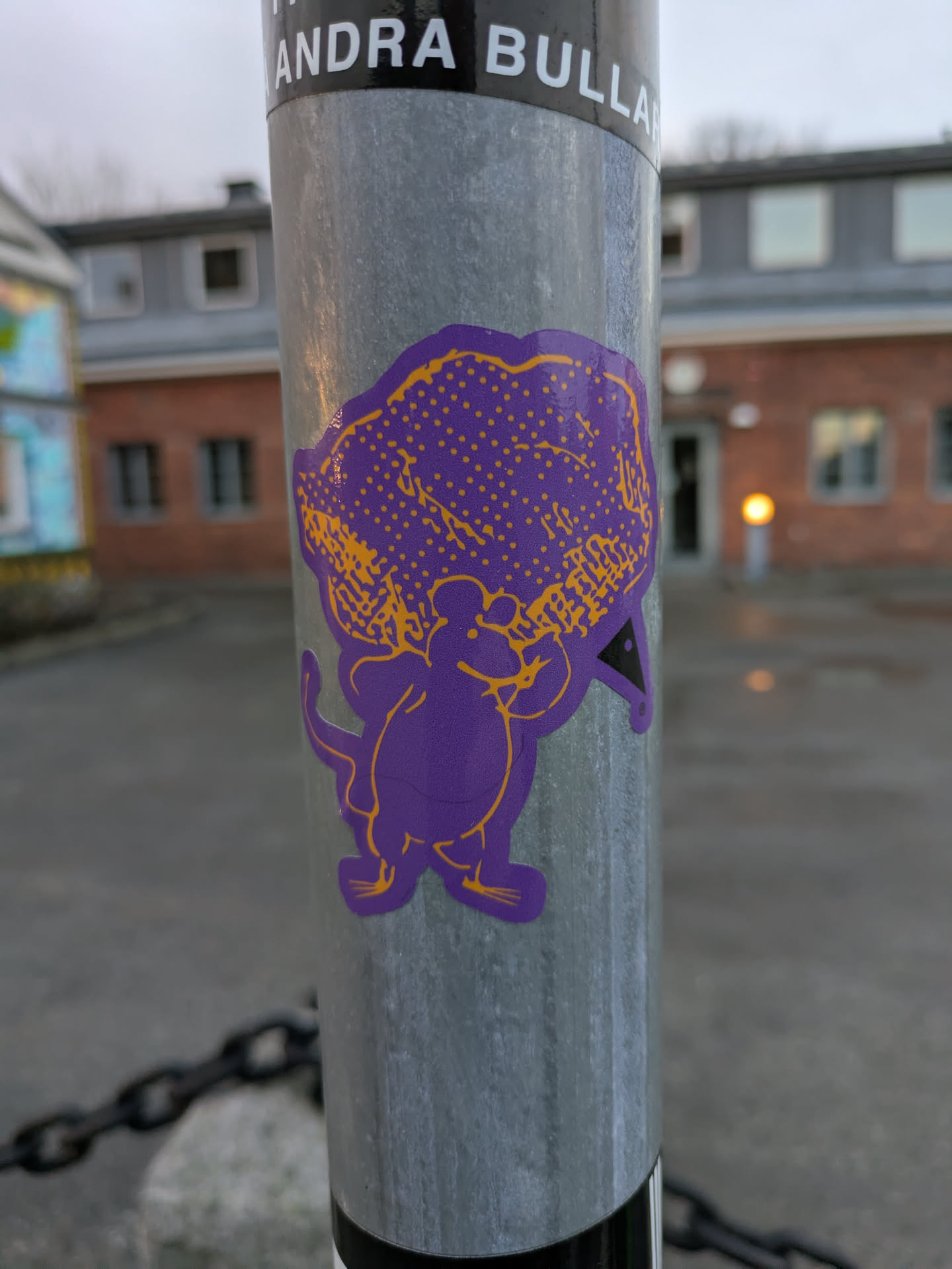 A close-up shot of a purple sticker featuring a mouse-like character with a large head and a small body. The character is depicted in a simple style with orange outlines. The sticker is affixed to a metal pole.