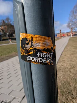 The sticker is rectangular with intentionally torn or distressed edges, giving it a rugged appearance. Its primary colors are a bright, almost neon orange and stark black, with white text. The background is predominantly orange, overlaid with a black, grungy, and splattered texture that suggests dirt or struggle. On the left side, a black silhouette of a person is depicted in the act of climbing over a fence or barrier, which is rendered with a mesh pattern and barbed wire elements in black against the orange. The right side of the sticker features bold, white, sans-serif text that reads "FIGHT BORDERS", with "FIGHT" positioned above "BORDERS". The overall design is high-contrast and impactful, conveying a strong message.