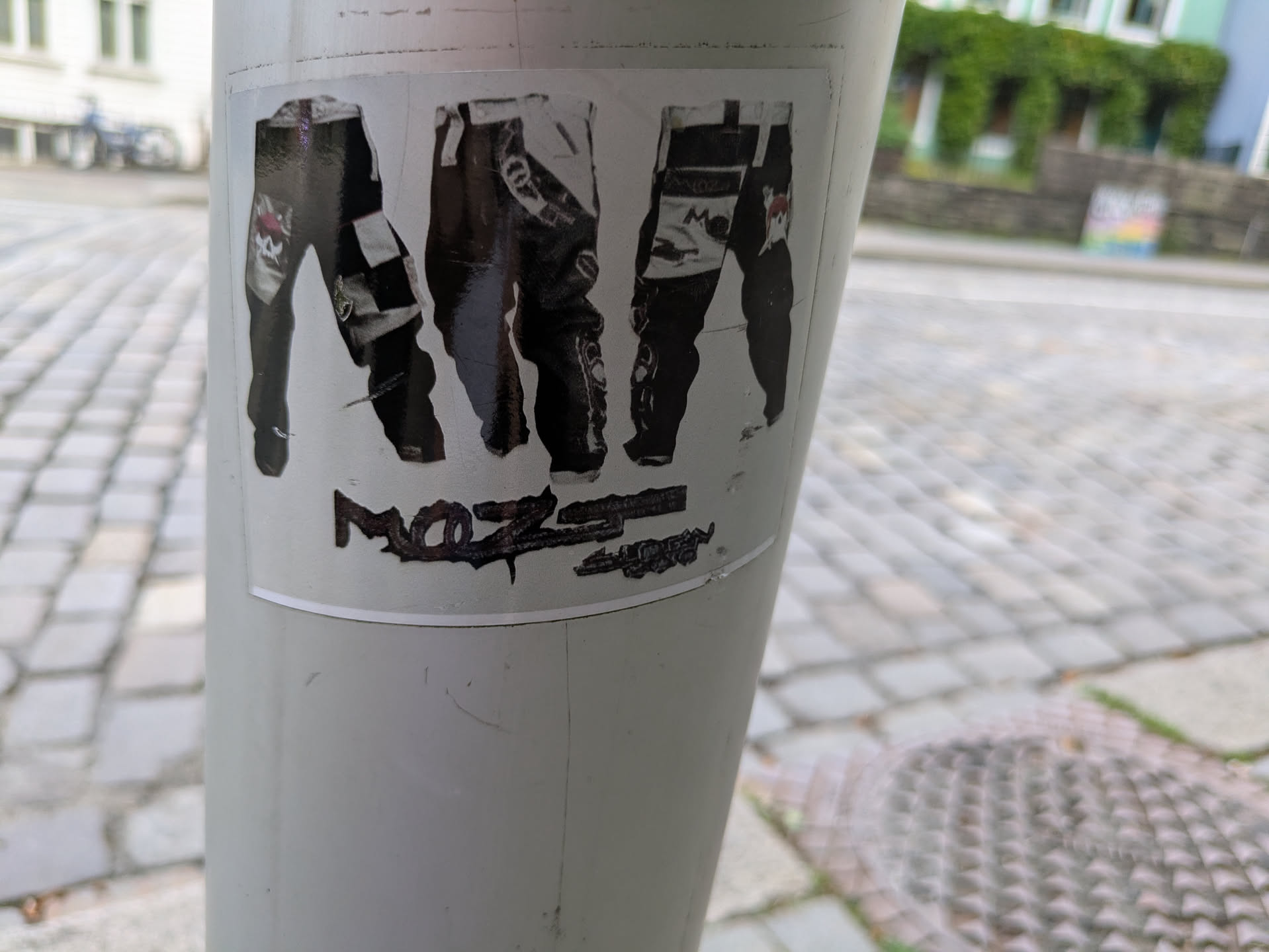 The sticker displays three separate images of what appear to be motocross or racing pants, each with distinct designs. The leftmost pair shows a black and white checkered pattern and a red graphic, possibly a skull or logo, near the knee. The middle pair is predominantly dark with some lighter accents, and the rightmost pair also features a mix of dark and light sections with a small red detail. Beneath the pants, there is dark, distressed text; the first part is decipherable as 'MoZ', while the subsequent text is heavily worn and largely illegible. The sticker is applied to a light-colored utility pole set against a background of cobblestone pavement and distant buildings.