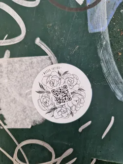 A round sticker featuring a line drawing of flowers surrounding a QR code.  The text 'Musa Tattoo' is subtly written at the top.