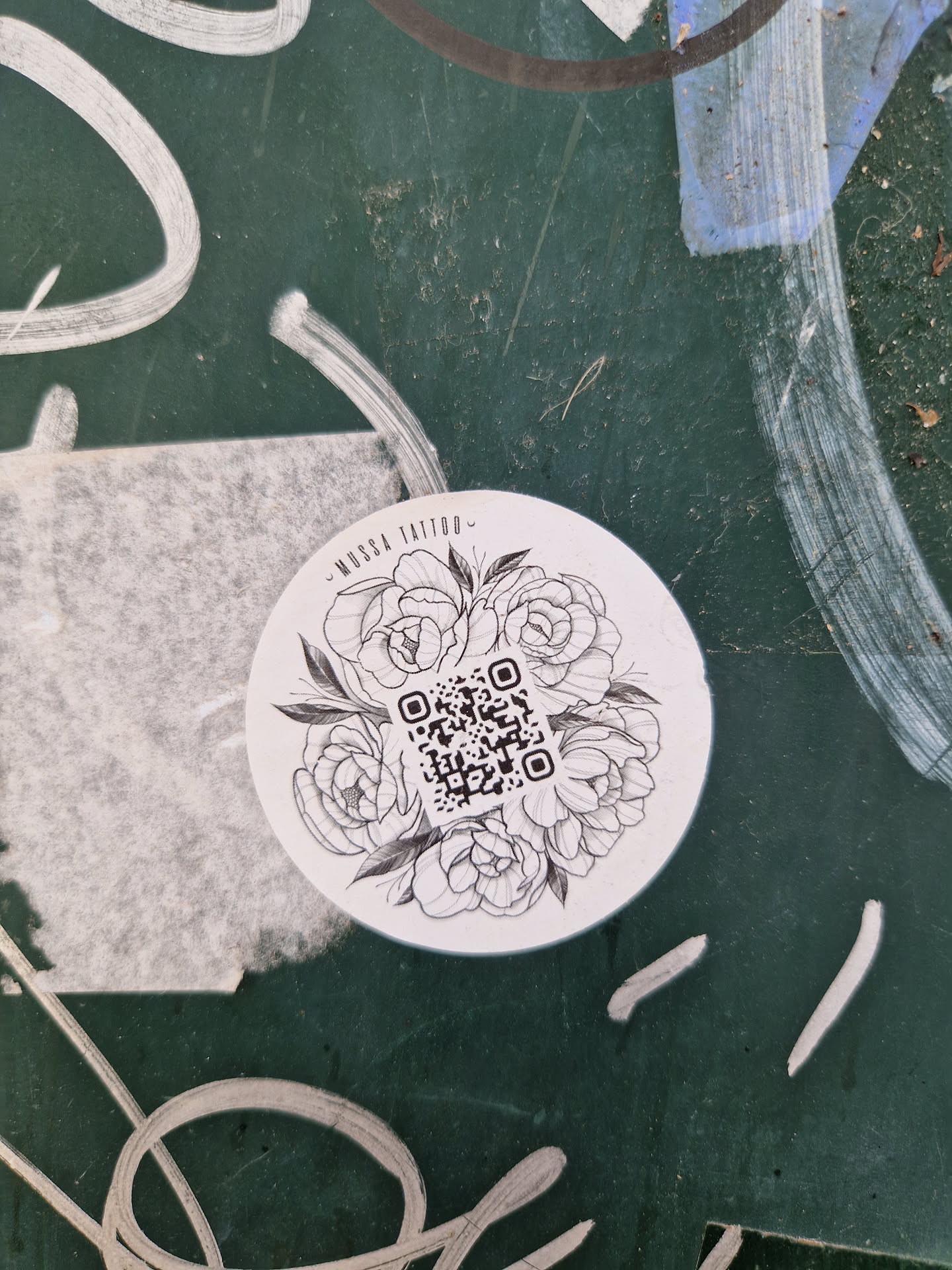 A round sticker featuring a line drawing of flowers surrounding a QR code.  The text 'Musa Tattoo' is subtly written at the top.