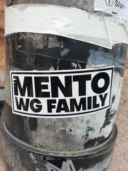 Black and white sticker with the text 'MENTO WG FAMILY' on it. The sticker is placed on a dirty, weathered surface.
