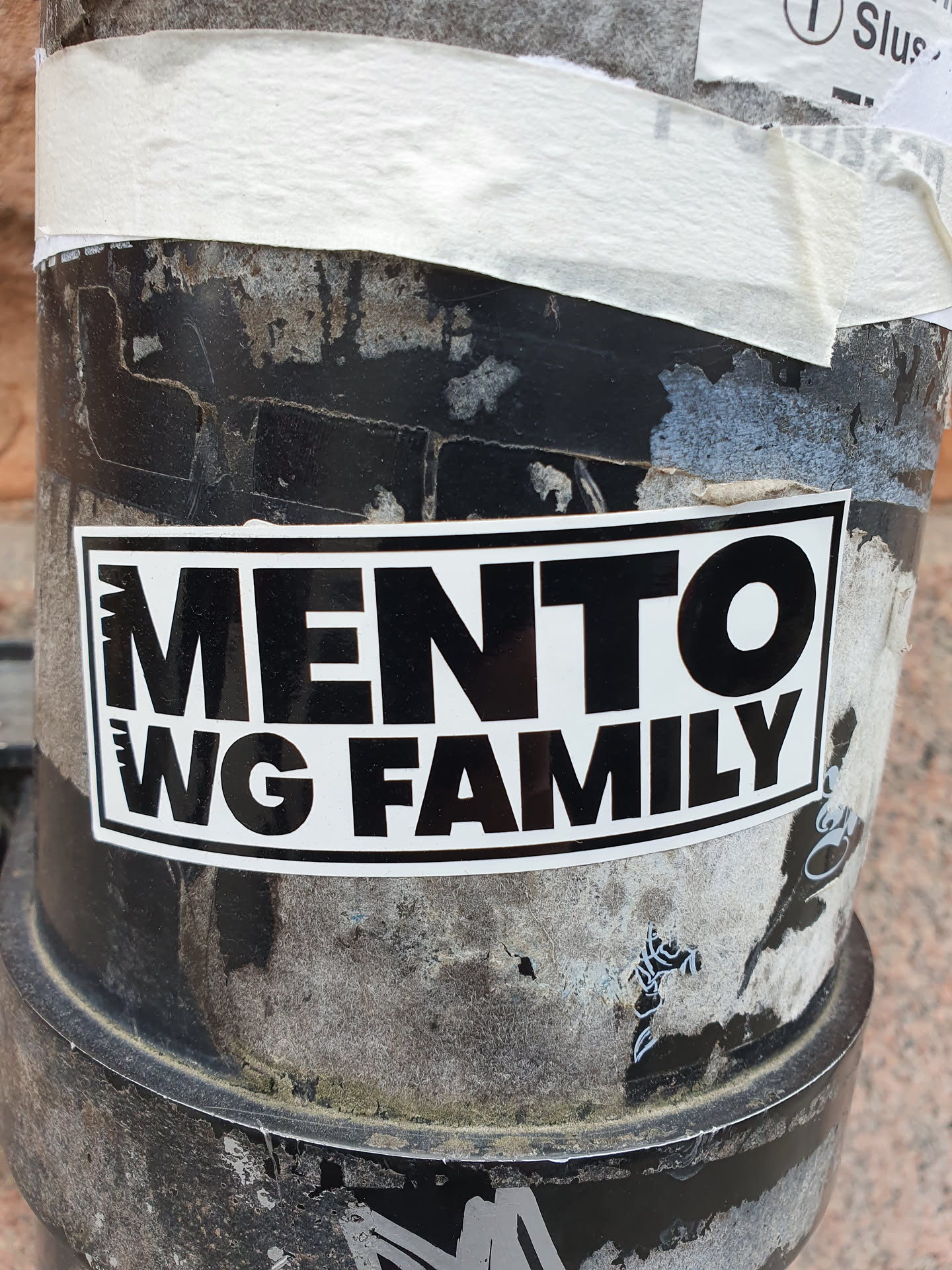 Black and white sticker with the text 'MENTO WG FAMILY' on it. The sticker is placed on a dirty, weathered surface.