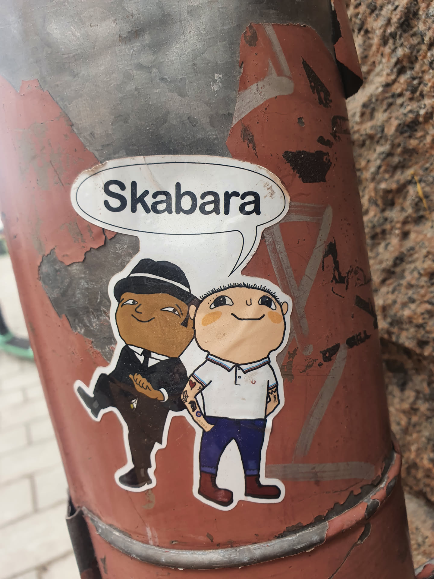 A sticker depicting two cartoon characters. One is wearing a suit and hat, and the other is wearing a polo shirt and jeans. A speech bubble above them says "Skabara". The sticker is applied to a weathered metal pole.
