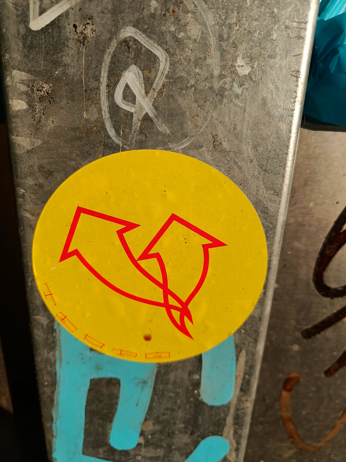 A round yellow sticker with two red upward arrows. The arrows are intertwined. The sticker is attached to a dirty gray metal surface.