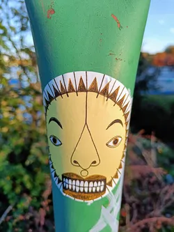 Close-up view of a round sticker featuring a stylized sun face with a prominent nose, expressive eyes, and a set of teeth. The sun has a jagged, radiating frame and the sticker is applied to a weathered green pole outdoors. The background is blurred, showcasing autumnal foliage.