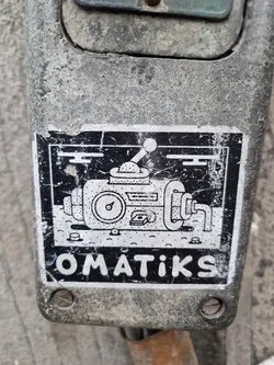 Black and white illustration of a machine with gauges and levers. The word OMATIKS is written at the bottom of the sticker. The sticker is affixed to a dirty, gray surface.