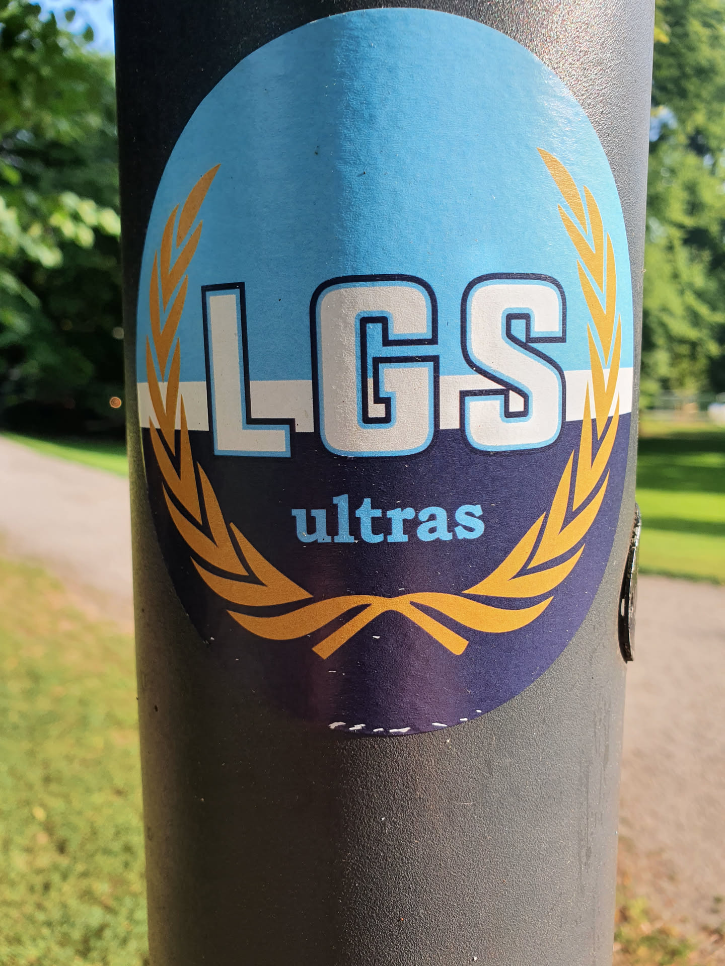 Oval sticker with the letters LGS in the center, surrounded by a golden laurel wreath. Below the letters, the word "ultras" is written in a smaller font. The background is blue, and the laurel wreath and letters are in white and gold. The sticker is applied to a dark gray pole.