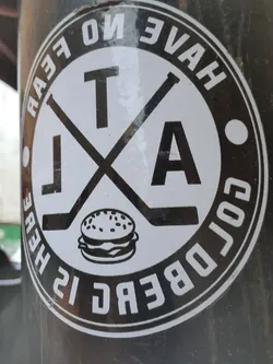 A black and white sticker with a logo. The logo includes crossed hockey sticks, the letters TA, a burger and the text "Goldberg is here. Have no fear."