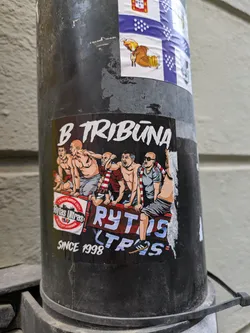 A sticker depicting a group of shirtless men sitting on a banner that says Rytas Ultras. The text B tribūna is written above the illustration.  The words Rytas Ultras and Since 1998 are also visible on the sticker. The sticker shows a group of fans, likely from a sports team.