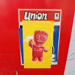 A sticker featuring a Haribo gummy bear wearing a 1. FC Union Berlin jersey. The gummy bear is holding a flare and has a happy expression. The background is yellow.