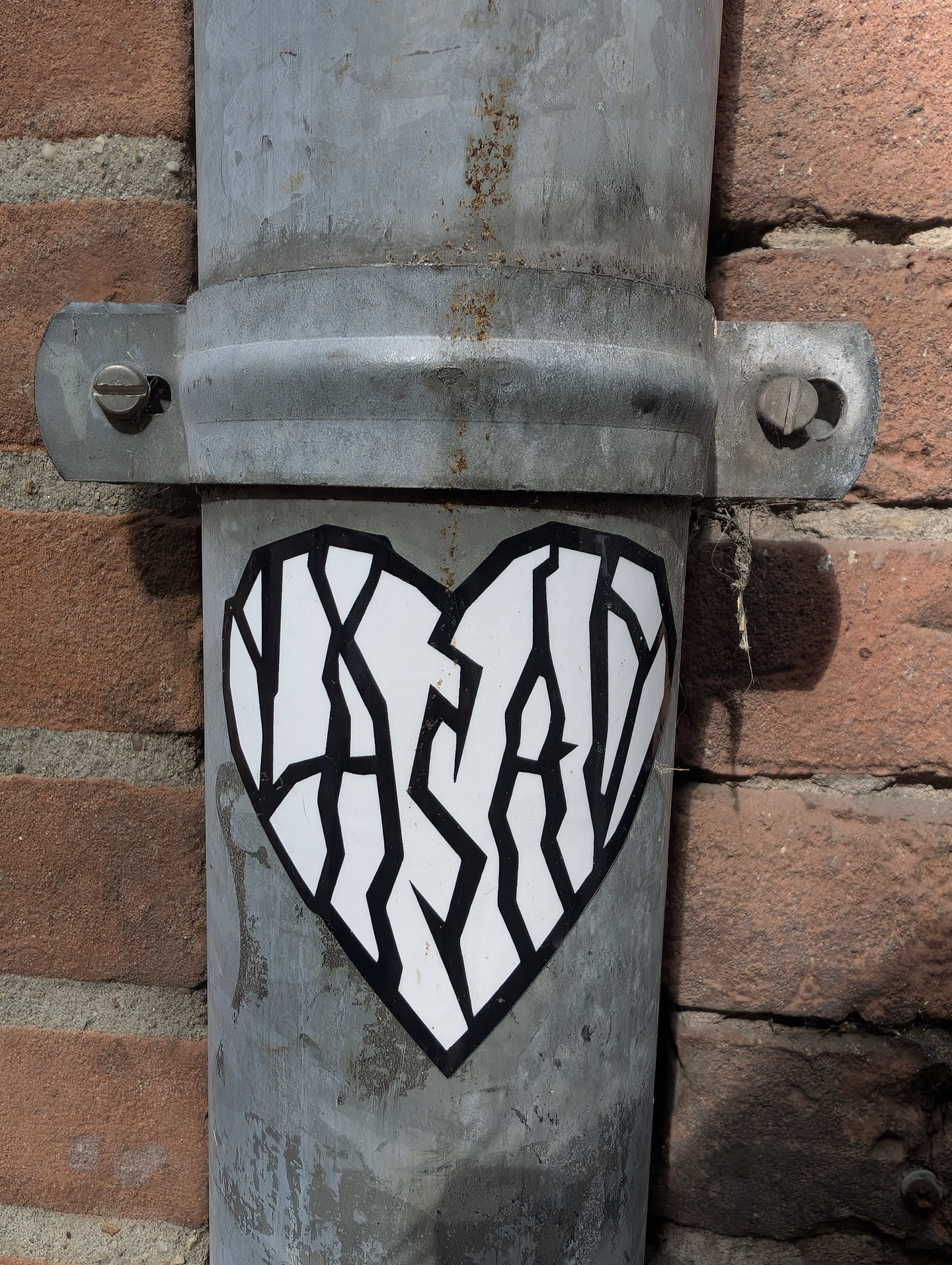 A white heart-shaped sticker with a black outline is placed on a gray metal pipe. Inside the heart, the text 'YASAY' is written in a stylized, fragmented font. The pipe is attached to a brick wall.