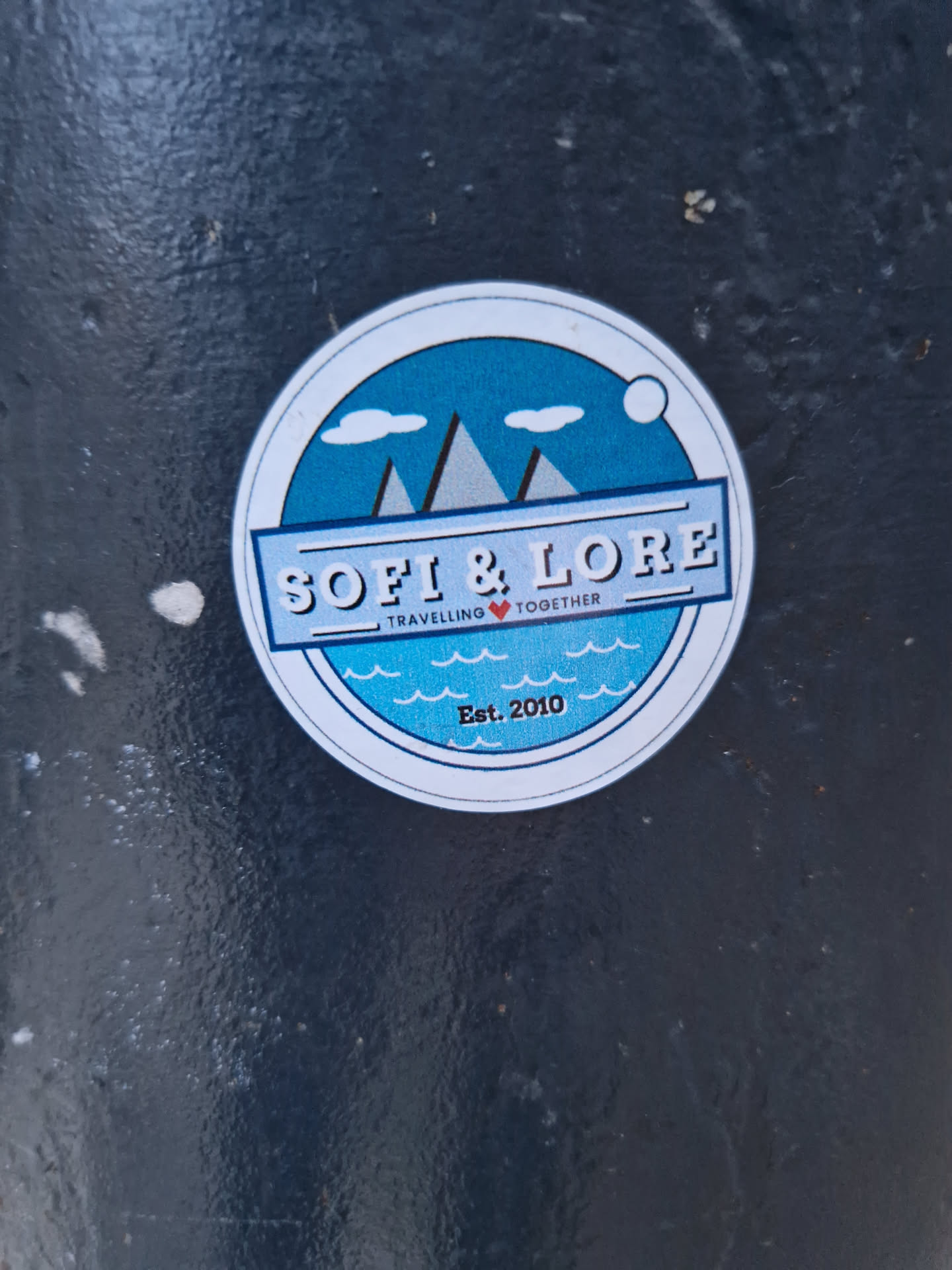 A round sticker with a blue design featuring mountains and waves.  The text 'SOFI & LORE' is prominently displayed, along with the tagline 'Travelling ❤️ Together' and 'Est. 2010'.