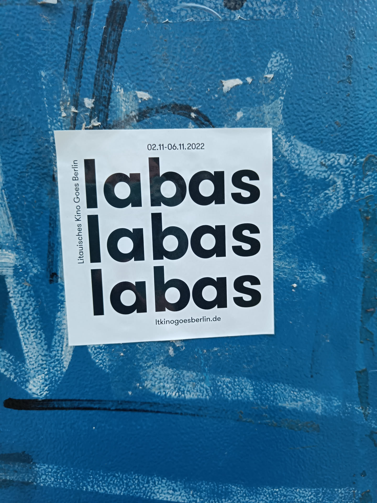 A minimalist sticker with the word "Labas" repeated three times.  The sticker also includes dates 02.11-06.11.2022 and a website address. It's stuck on a blue wall.