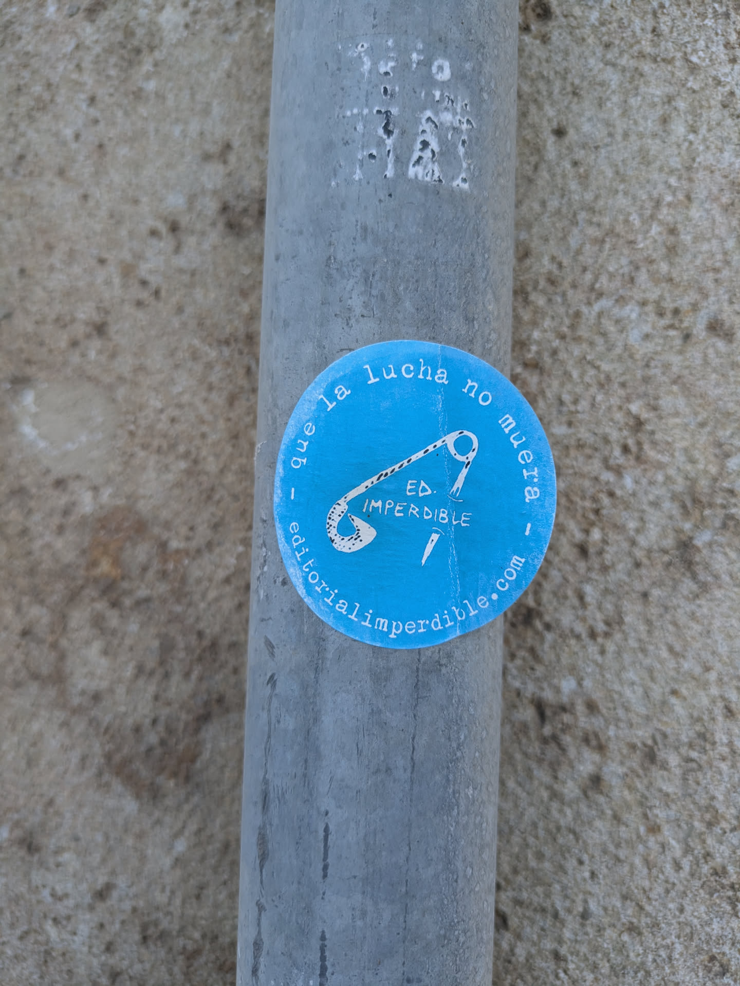 The sticker is a bright blue circle with white text around the border that reads "que la lucha no muera - editorialimperdible.com". In the center is a drawing of a needle and thread and under it, the text "ED. IMPERDIBLE".