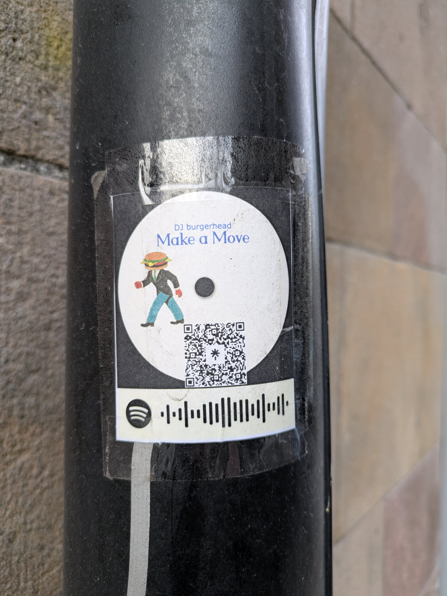 A round sticker featuring a burger-headed man in a suit walking. Above the figure is written "DJ burgerhead" and "Make a Move." Below the character, there is a QR code and a Spotify code.