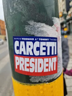 A sticker featuring a presidential campaign design for Thomas J. "Tommy" Carcetti. The sticker is affixed to a green pole and shows some wear and tear.