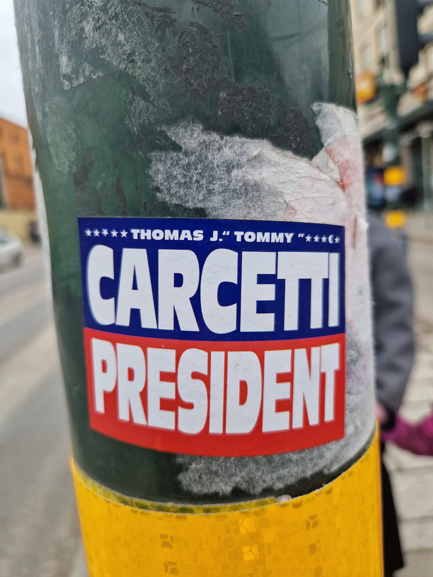 A sticker featuring a presidential campaign design for Thomas J. "Tommy" Carcetti. The sticker is affixed to a green pole and shows some wear and tear.