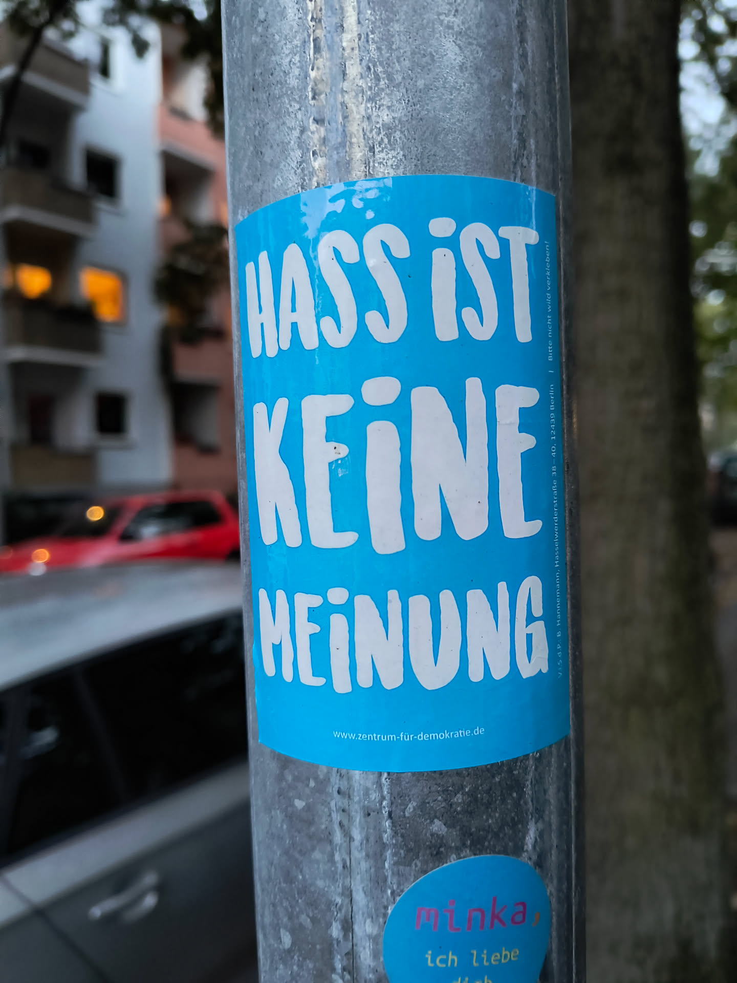 The central sticker is rectangular with rounded corners and a bright blue background. It features large, white, sans-serif lettering that reads 'HASS IST KEINE MEINUNG' (Hate is not an opinion). Below this main text, in smaller white font, is the website 'www.zentrum-fur-demokratie.de'. To the right of the main text, in a vertical orientation and very small font, is additional text that appears to be contact or legal information. There is also a smaller, oval-shaped sticker at the bottom right of the main sticker, with pink and yellow text on a blue background, which reads 'minka, ich liebe dich'.