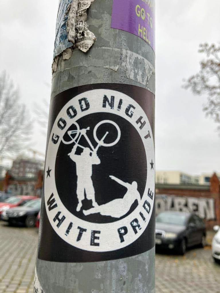 A circular sticker with a stark black background and a white outer border. Inside the black circle, two stylized white figures are depicted. One figure stands upright, holding a bicycle overhead, seemingly in an act of aggression or confrontation towards a second figure that lies prone on the ground. The phrase 'GOOD NIGHT' is arched in white uppercase letters across the top inner part of the white border, and 'WHITE PRIDE' arches along the bottom. Small white stars are positioned on either side, separating the top and bottom text from the central imagery.