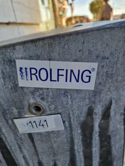 A close-up shot of a Rolfing sticker affixed to a weathered gray surface. The sticker is white with blue text and a small graphic.  There is also a smaller, white label with the number 1141 visible below the sticker.