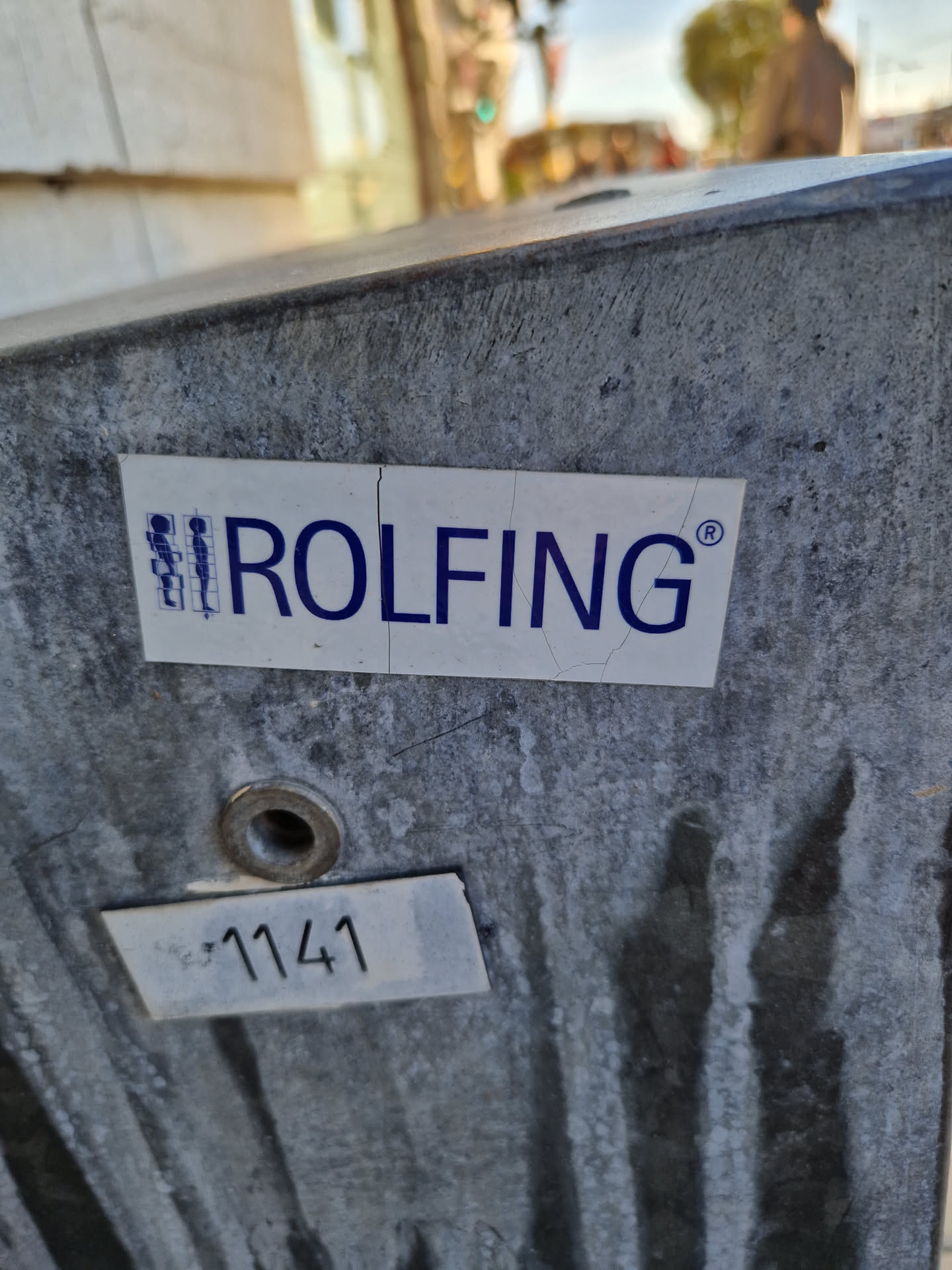 A close-up shot of a Rolfing sticker affixed to a weathered gray surface. The sticker is white with blue text and a small graphic.  There is also a smaller, white label with the number 1141 visible below the sticker.