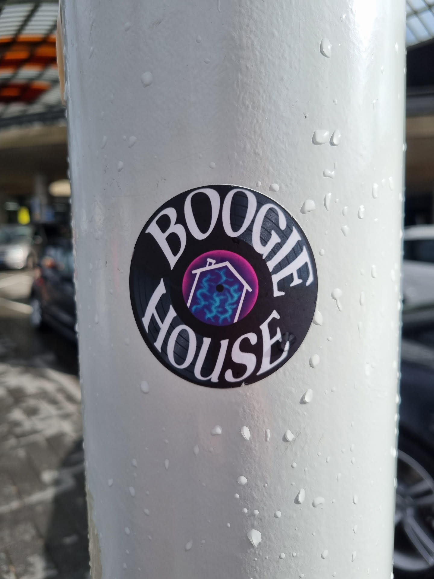 A round sticker featuring the text 'BOOGIE HOUSE' in a stylized font reminiscent of vinyl records.  Inside the circle is a smaller image of a house with purple and blue lightning bolt designs. The sticker is affixed to a light gray pole outdoors, and there are water droplets visible on the pole's surface.