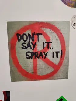 Sticker depicting graffiti art on a brick wall. The graffiti is a red circle with a peace symbol inside, with the words "Don't say it... Spray it!" written in black paint.