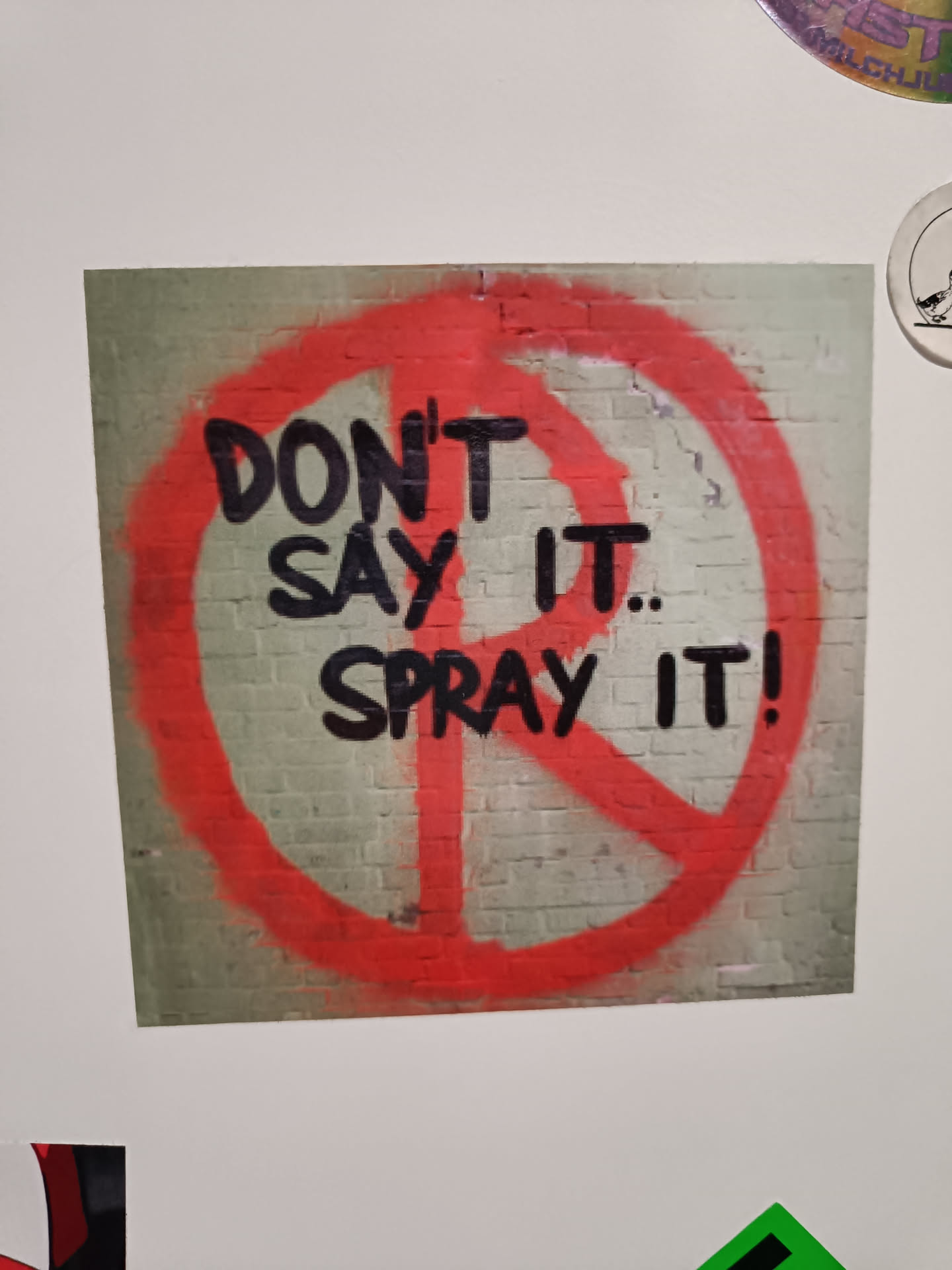 Sticker depicting graffiti art on a brick wall. The graffiti is a red circle with a peace symbol inside, with the words "Don't say it... Spray it!" written in black paint.