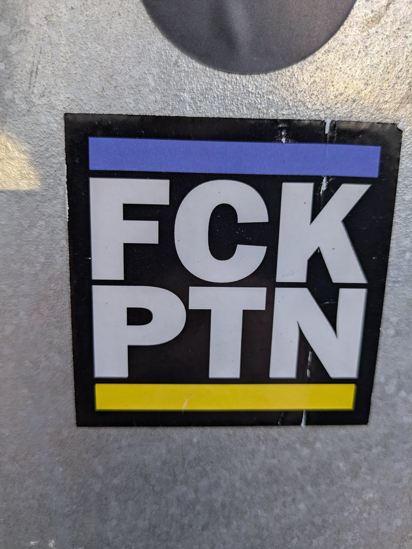 A black sticker with white lettering that says "FC*PTN". It has a thin yellow stripe at the bottom and a thin light blue stripe at the top.