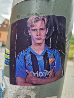 A sticker featuring a young man with blond hair, wearing a blue and black soccer jersey. He has his arms crossed and appears confident. The jersey has various sponsors' logos, including 'Prioritet' and 'Adidas'. The sticker is affixed to a weathered pole, adding to its urban aesthetic.