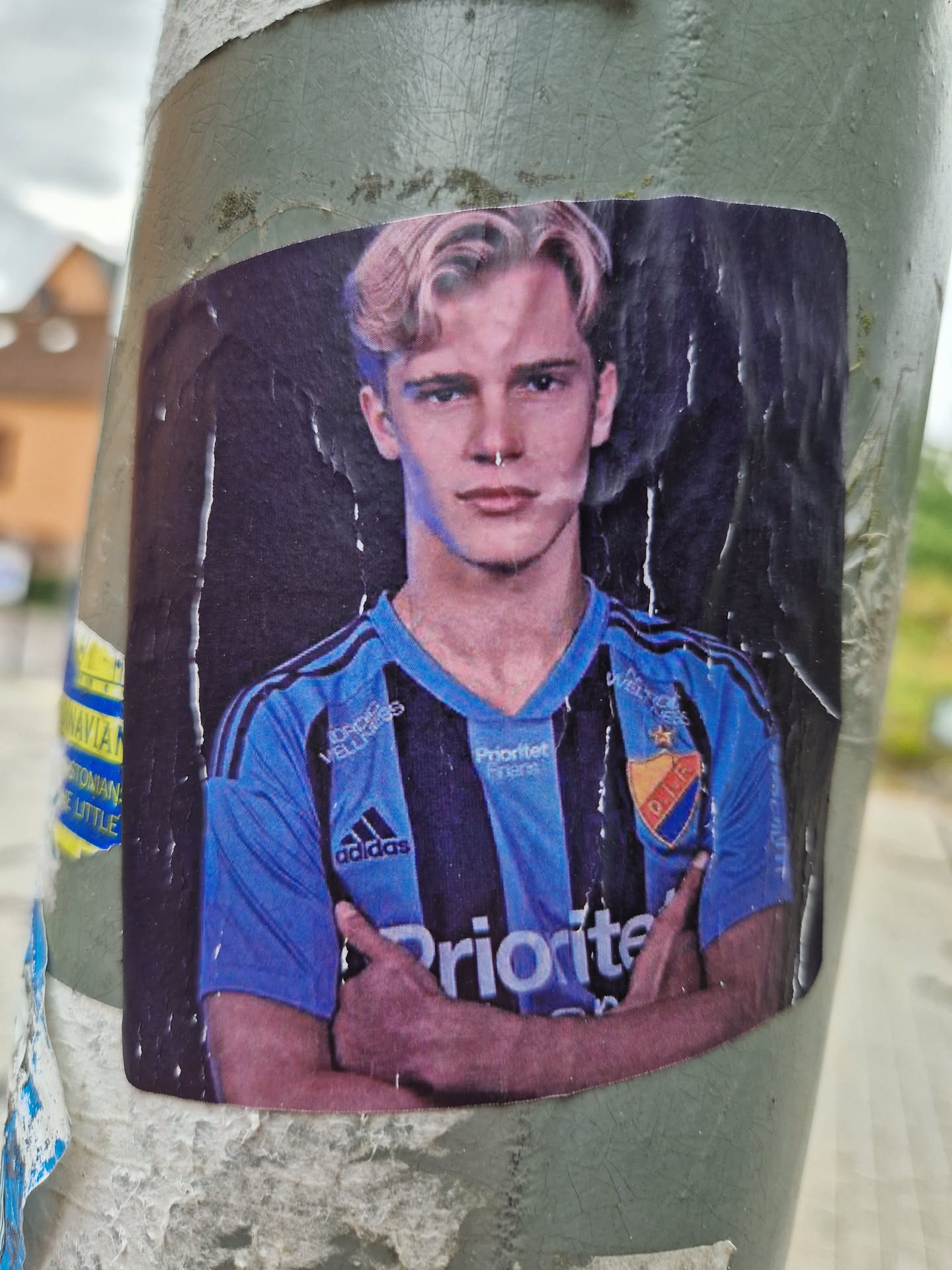 A sticker featuring a young man with blond hair, wearing a blue and black soccer jersey. He has his arms crossed and appears confident. The jersey has various sponsors' logos, including 'Prioritet' and 'Adidas'. The sticker is affixed to a weathered pole, adding to its urban aesthetic.