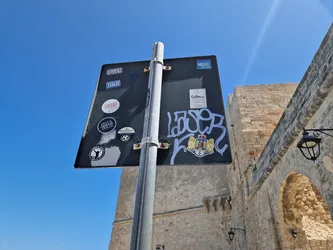 A collection of stickers adorns a sign, including a prominent crest-like sticker, possibly representing a local symbol or organization. The sign also features some graffiti. The background shows an old stone building, possibly in a European city. Several other stickers are visible around it, with text mostly unreadable.
