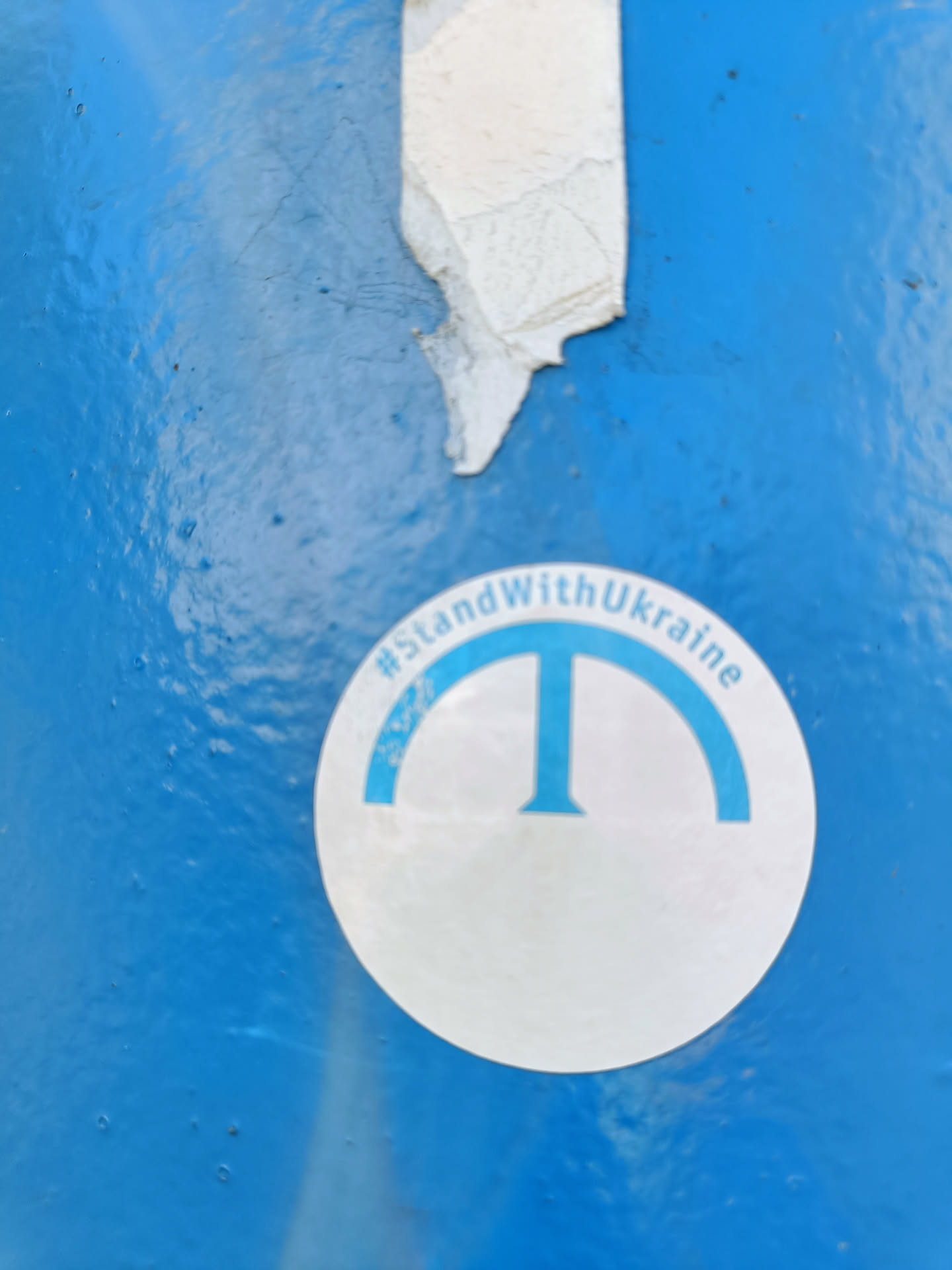A round white sticker on a blue surface with a light blue 'T' symbol and the text '#StandWithUkraine' around it.