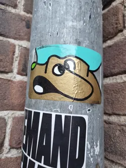 A gold-colored dog sticker with a teal hat. The dog has large eyes and a surprised expression.  It's a simple yet charming design.