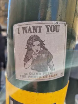 A sticker featuring a woman pointing at the viewer, similar to the iconic 'I Want You' poster. The text includes a call to stand with Iranian women.