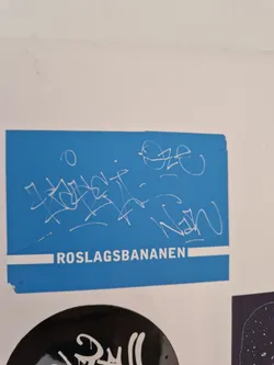 A blue sticker with white graffiti art and the text "ROSLAGSBANANEN" at the bottom. There are several tags on the sticker, including what looks like signatures.