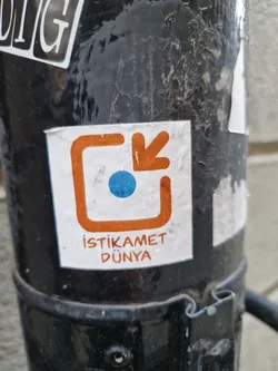 A sticker with an orange square and arrow pointing down, a blue dot inside. The text below says "İstikamet Dünya", which translates to "Direction World" in Turkish.