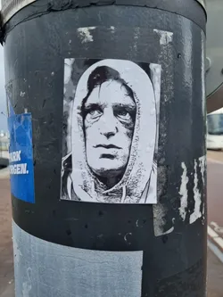 A grayscale sticker featuring a stylized portrait of a man with a hooded sweatshirt. The image has a slightly gritty texture, adding to its artistic feel. The sticker is affixed to a dark gray pole, which shows signs of wear and other stickers.