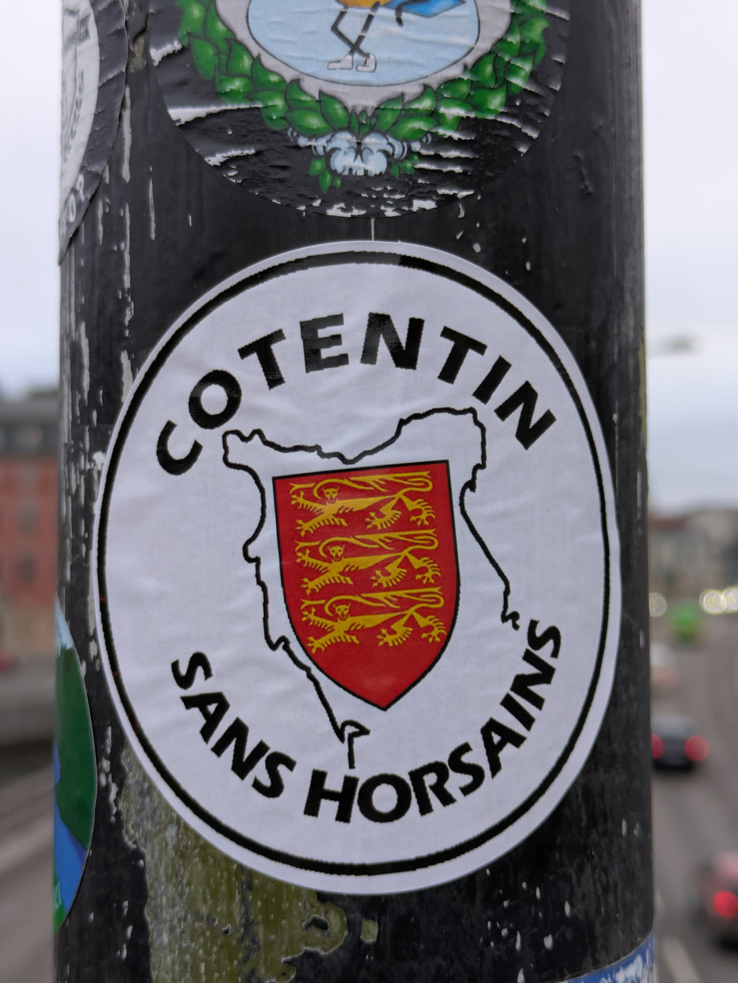 A round sticker with the text "COTENTIN" at the top and "SANS HORSAINS" at the bottom.  In between is an outline of the Cotentin Peninsula with a coat of arms featuring golden lions on a red shield. The sticker is affixed to a dark-colored pole.