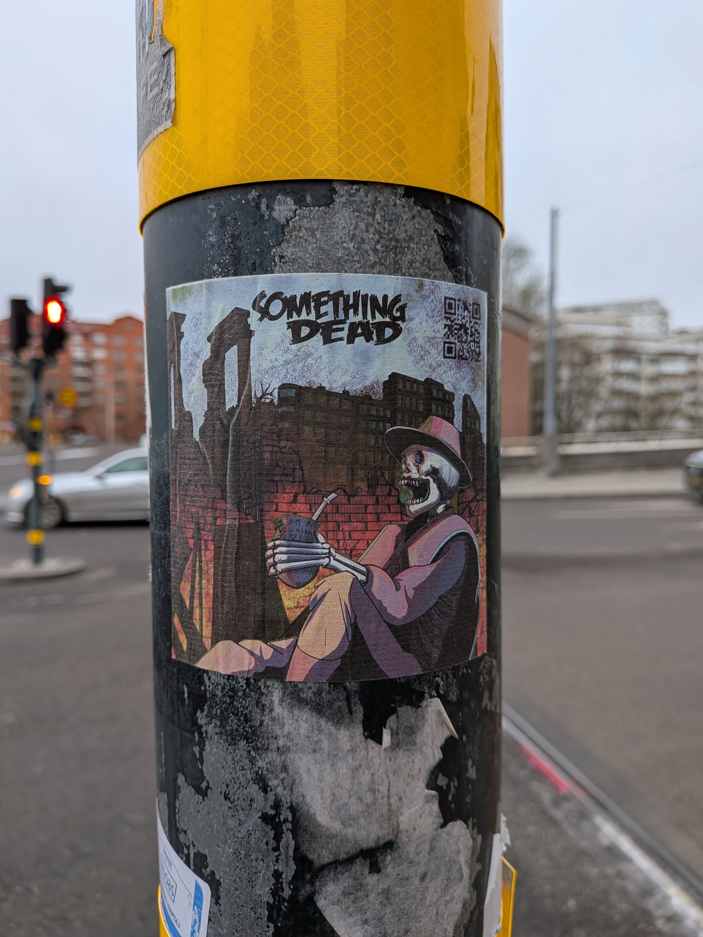 The sticker features a stylized illustration of a skeleton wearing a wide-brimmed hat and a cloak. The skeleton is seated and holding a gourd with a straw, appearing to drink. The background depicts a desolate urban landscape with crumbling brick buildings and structures, rendered in muted tones of brown, red, and grey. The text 'SOMETHING DEAD' is prominently displayed in a distressed, bold font above the scene. A QR code is visible to the right of the text.