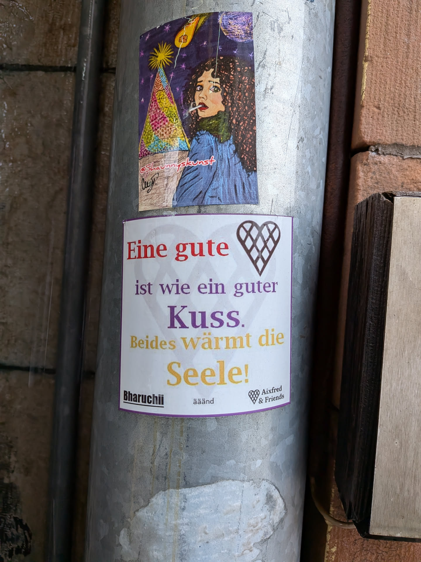 The sticker is a white, rectangular print with a thin purple border along its bottom edge. It features a German quote in varying font sizes and and colors. In the top right corner, there is a brown, stylized heart icon with a grid pattern inside. At the bottom, below the quote, are the names 'Bharuchii' and 'Aixfred & Friends', separated by 'ääänd', and another smaller, similar heart icon next to 'Aixfred & Friends'. The text colors are red, grey/purple, purple, and yellow/gold.