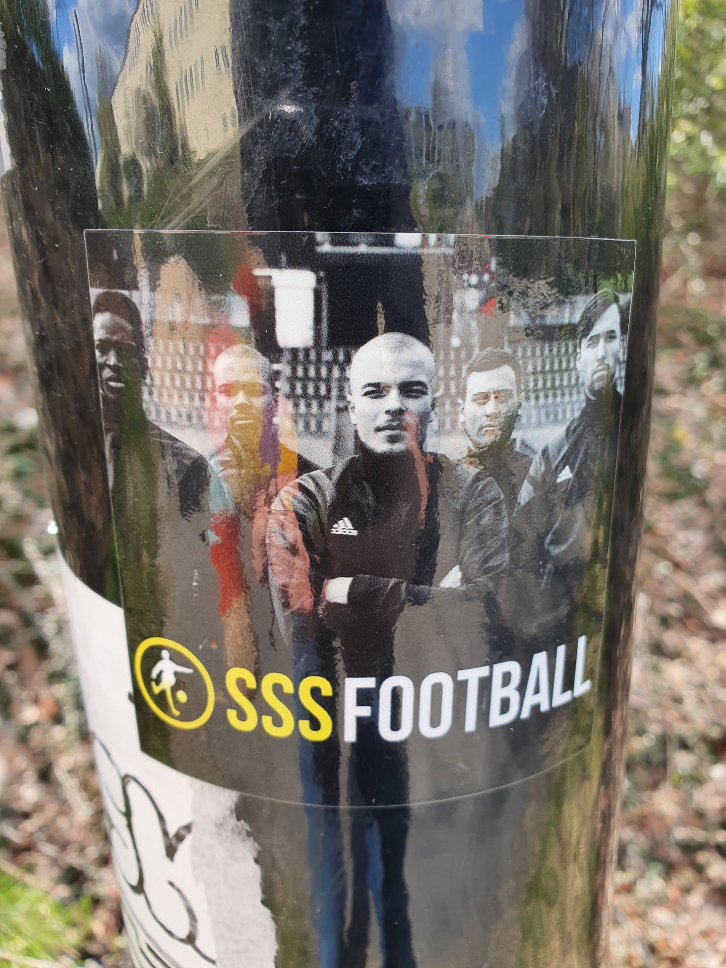 A black and white sticker featuring a group of men, possibly a football team, with the text 'SSSFOOTBALL' underneath. There's a small logo of a football player to the left of the text. The background image appears to be a street scene.