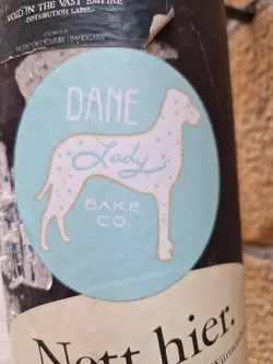 A light teal round sticker with a graphic of a Great Dane. The text on the sticker says "DANE Lady BAKE CO." in a simple, hand-drawn style font. The Great Dane is depicted in a light beige color with dark spots. The sticker has a slightly worn look.