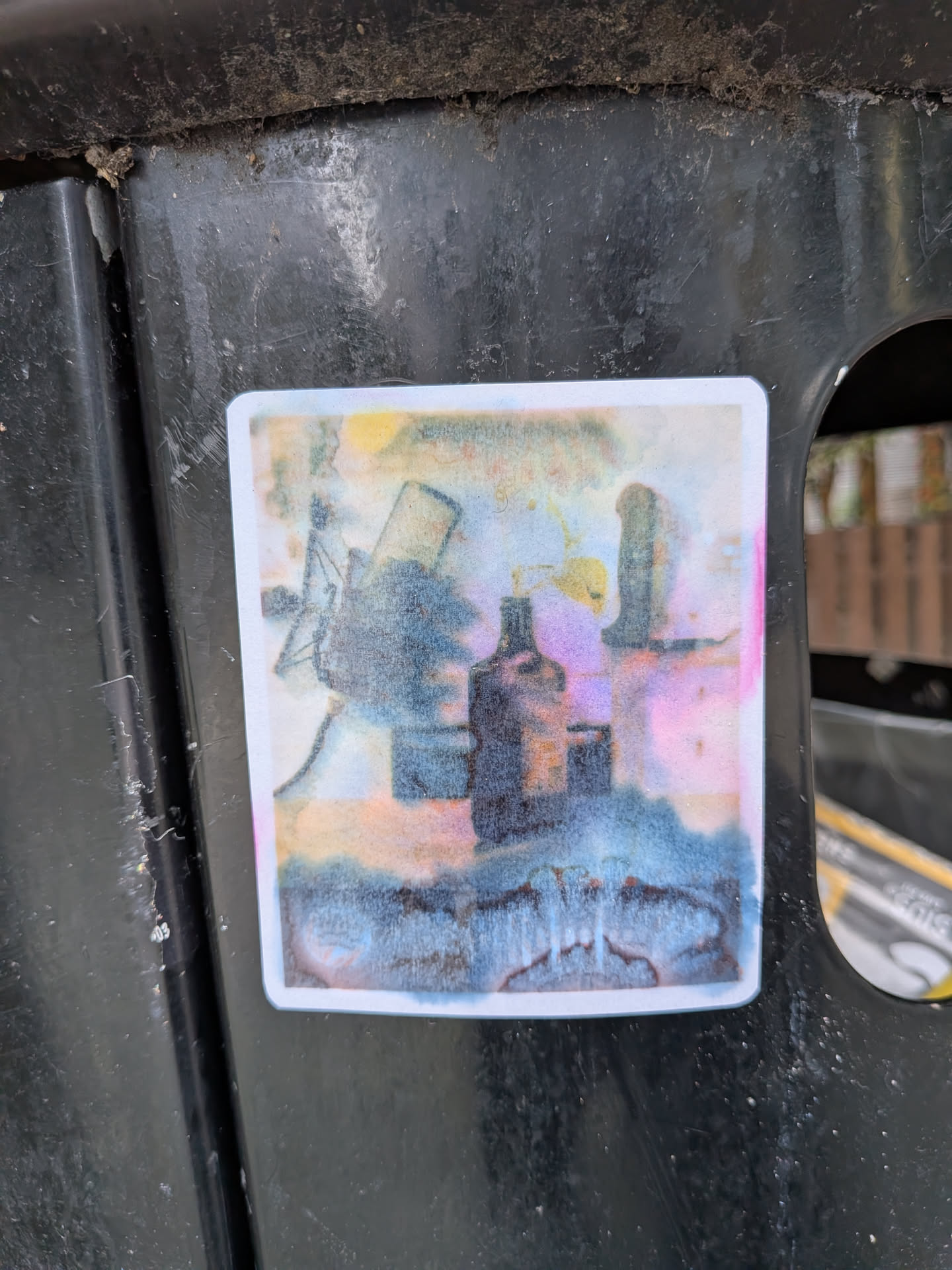 A rectangular sticker with a white border featuring a faded, watercolor-style image. It depicts a central bottle silhouette, a windmill structure on the left, and a tower on the right, rendered in bleeding blue, pink, and yellow tones.