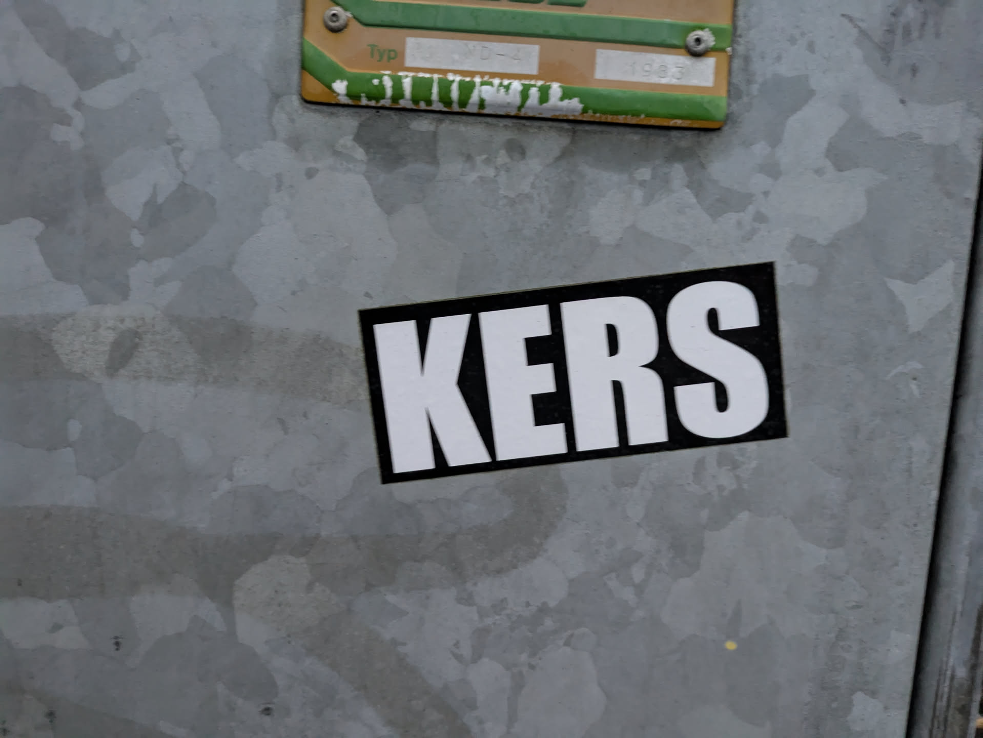 A close-up shot of a black and white sticker with the word "KERS" in bold, uppercase letters. The sticker is affixed to a gray metal surface with a textured finish.