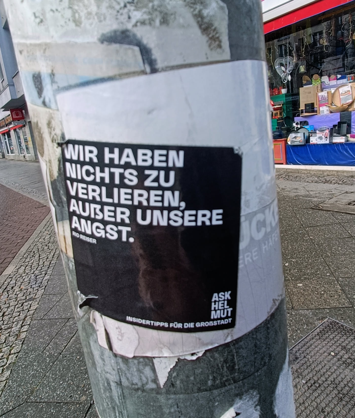 A black and white sticker on a pole in a city. The text says 'Wir haben nichts zu verlieren, außer unsere Angst.' which translates to 'We have nothing to lose, except our fear.'  There is also smaller text that says 'Insidertipps für die Großstadt' (Insider tips for the big city) and 'Ask Helmut Mut'