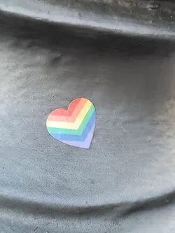 A small sticker in the shape of a heart with rainbow colors is affixed to a dark gray surface. The sticker appears to be made of vinyl or a similar material.