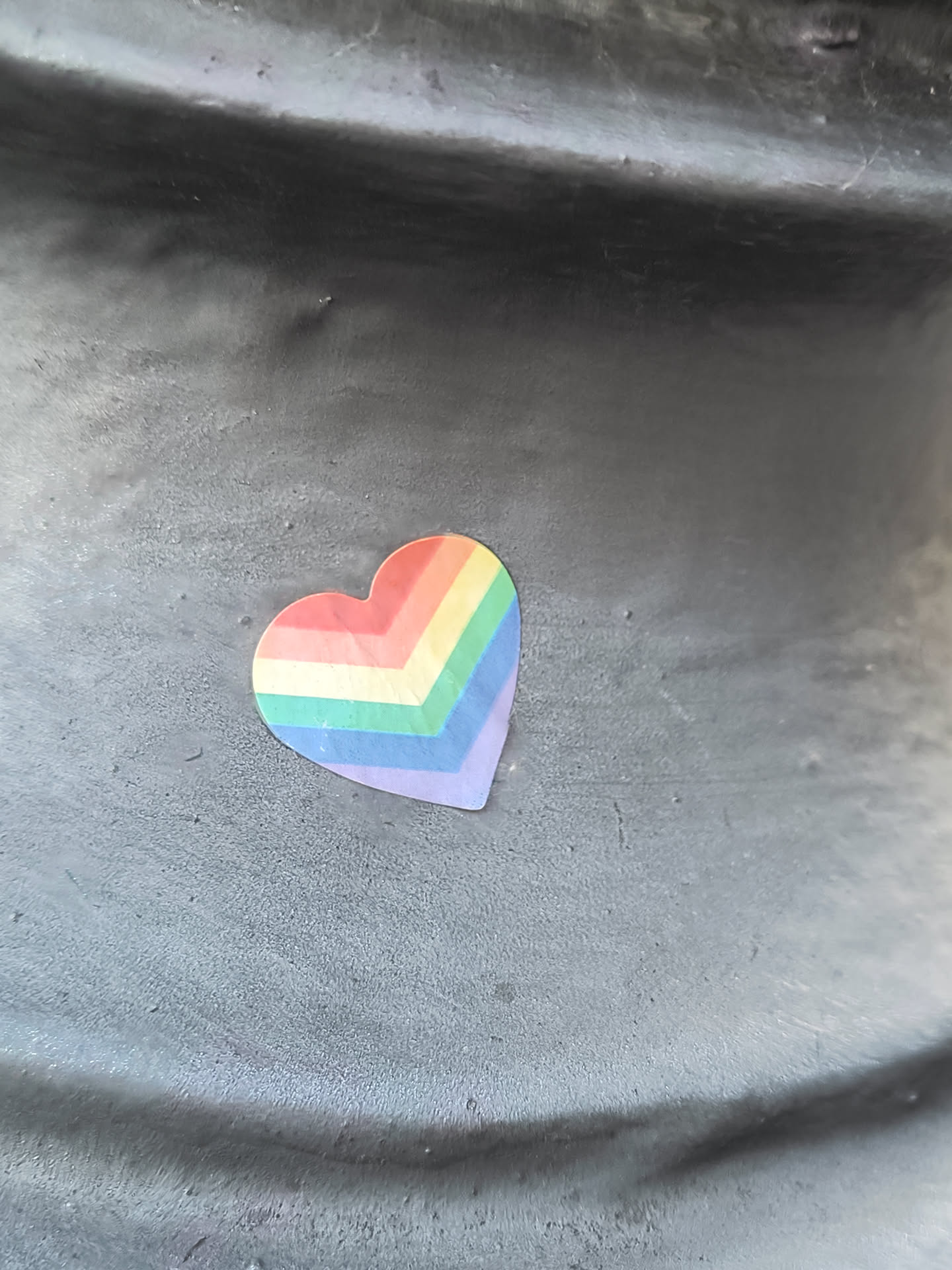 A small sticker in the shape of a heart with rainbow colors is affixed to a dark gray surface. The sticker appears to be made of vinyl or a similar material.